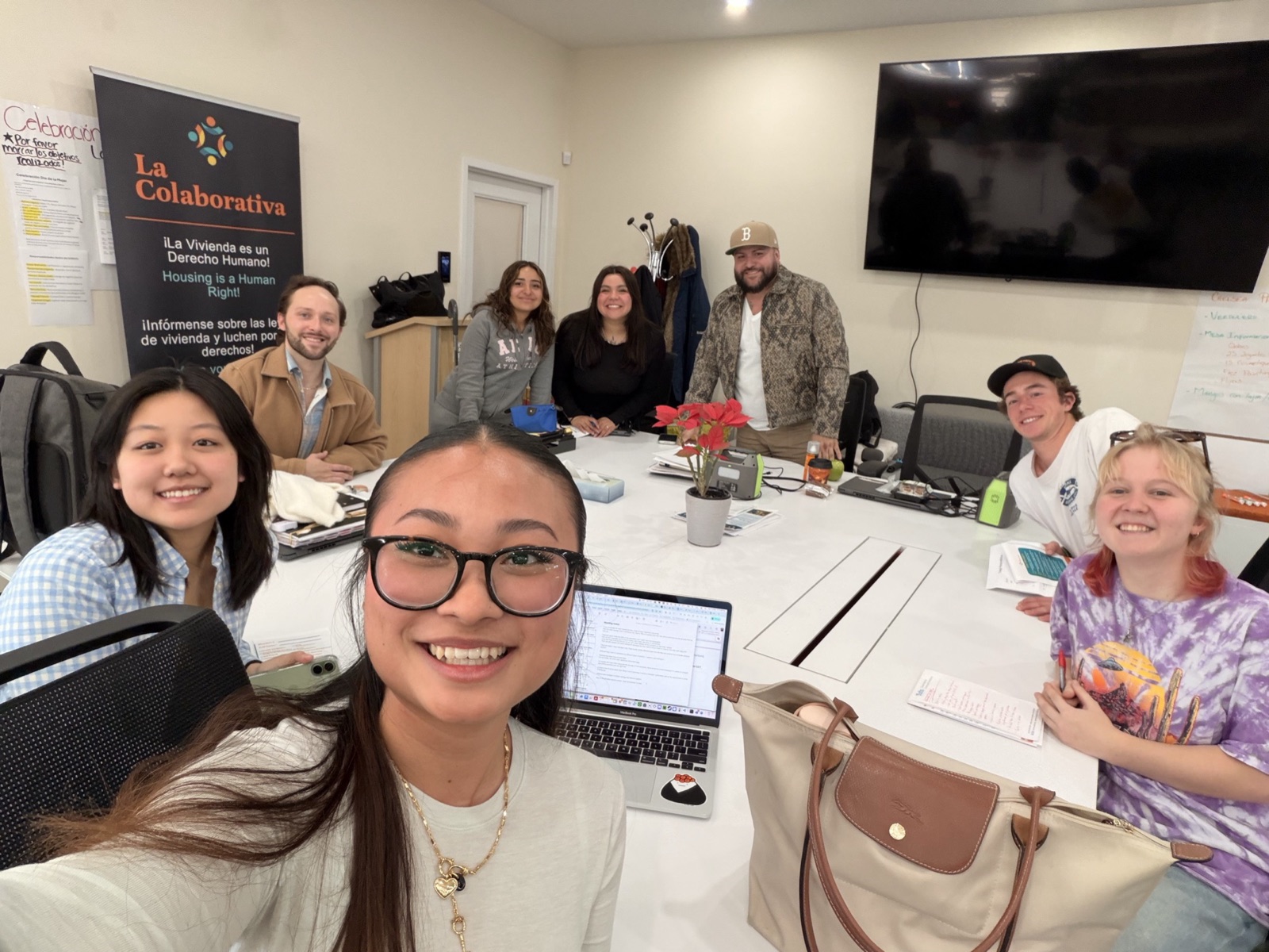 Vorleak taking a selfie at a conference table with the project team and La Colaborativa staff, a La Colaborativa banner reading "Housing is a Human Right" in the background